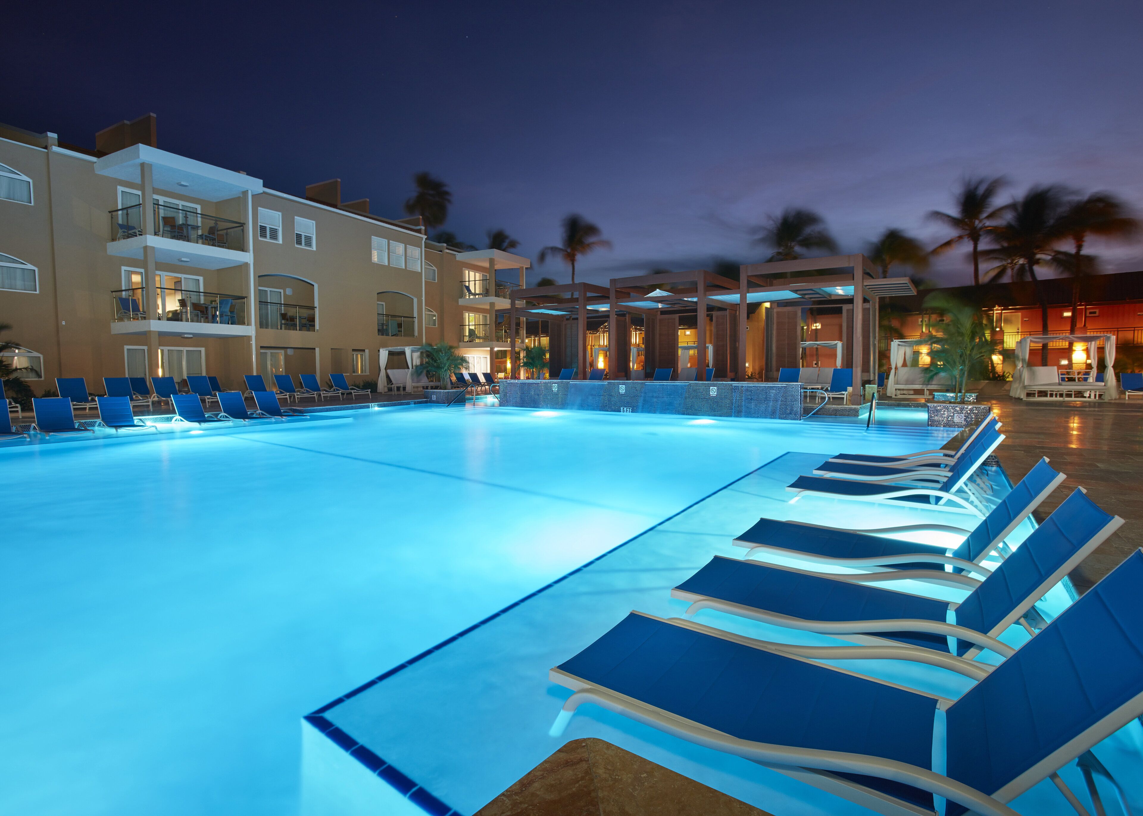 A swimming pool with lounge chairs, a covered seating area, and a building with multiple floors and balconies.