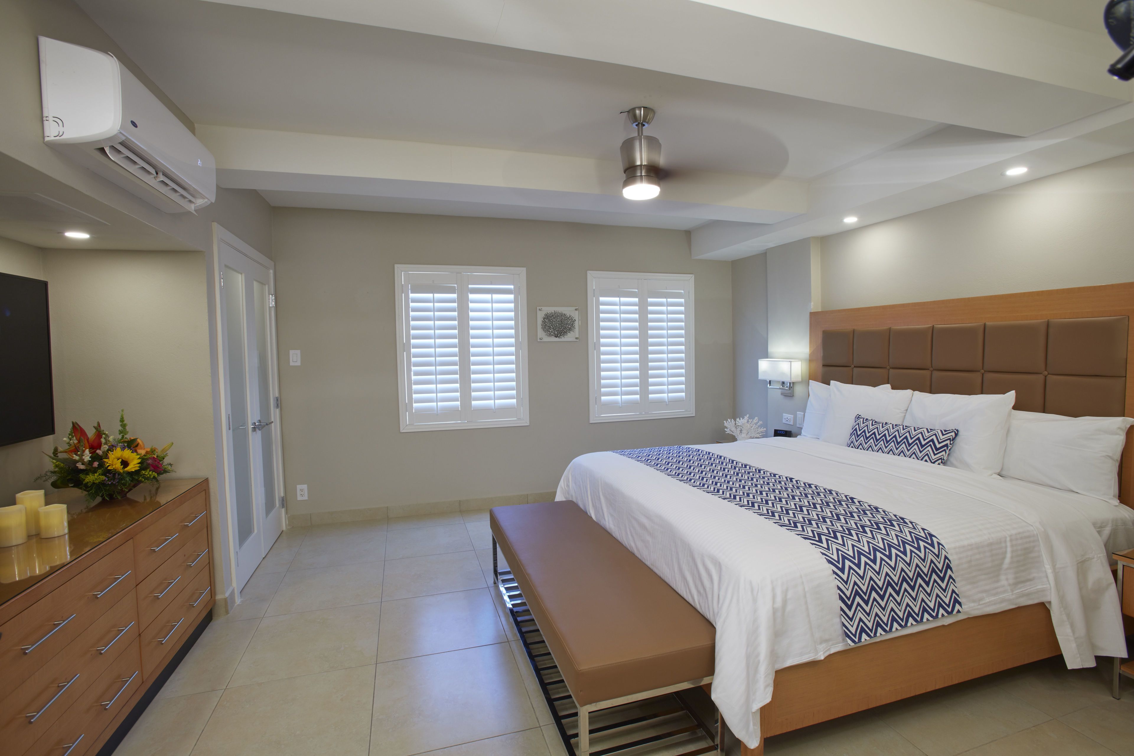 A bedroom with a bed, bench, dresser, TV, and air conditioning unit.