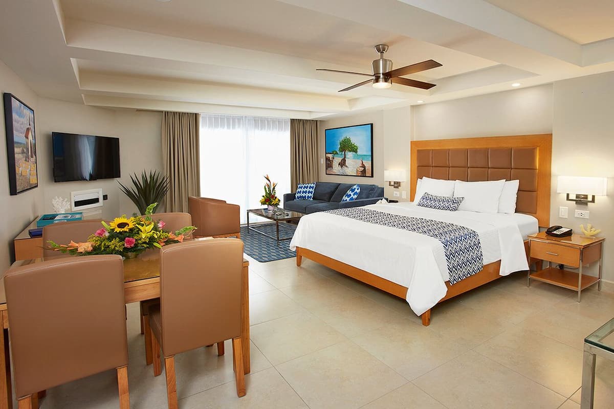 A hotel room with a bed, a desk with chairs, a television, a ceiling fan, and a painting on the wall.