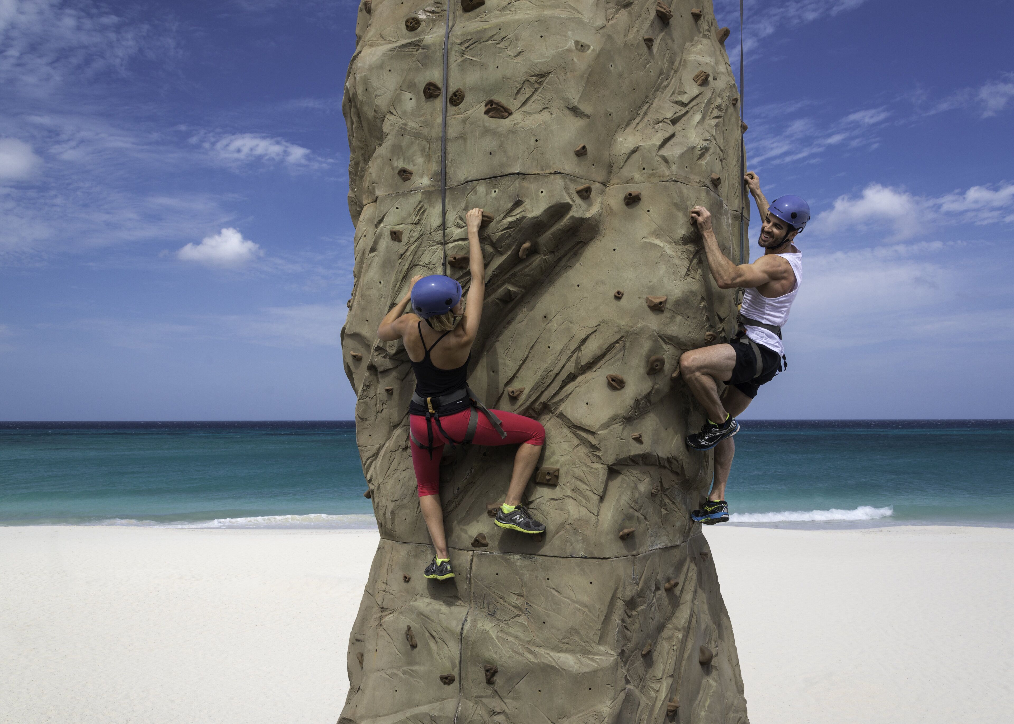 Two people rock climbing on a large rock formation by a beach with clear skies.