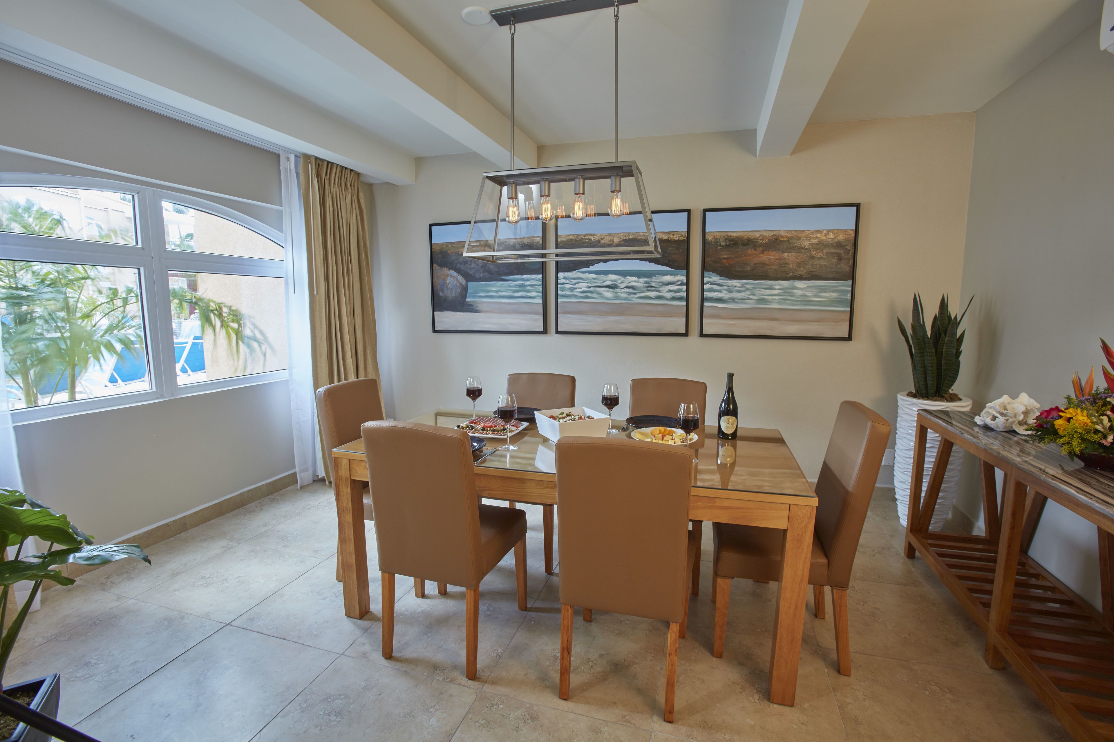 Un coin repas avec une table dressée pour six personnes, entourée de chaises, d’un luminaire suspendu et de deux tableaux encadrés accrochés au mur.