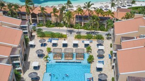 A resort with a central swimming pool surrounded by lounge chairs and umbrellas, adjacent to a beach with palm trees and a clear blue sea. - Divi Dutch Village Beach Resort (Oranjestad)