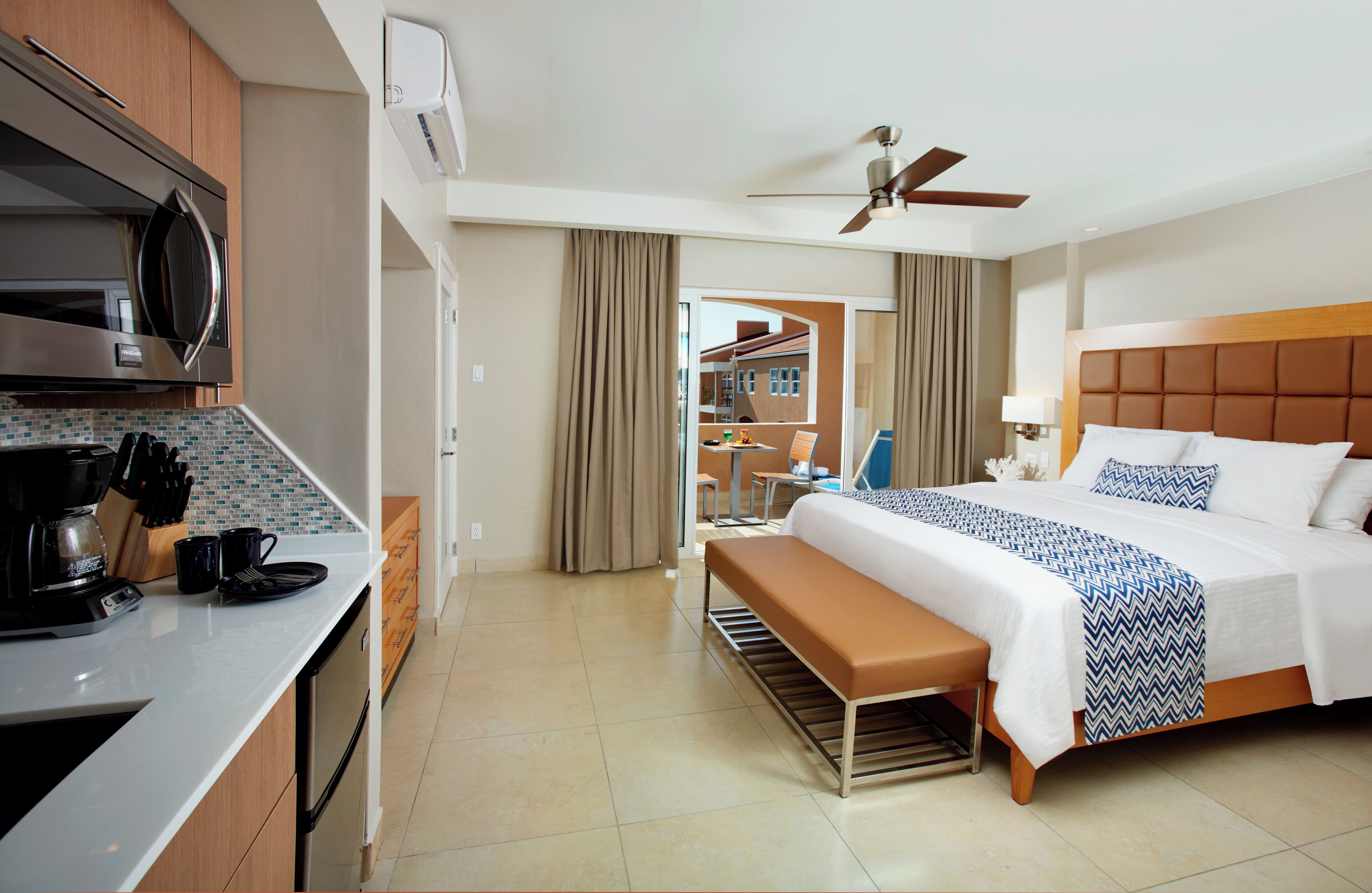 A hotel room with a bed, a bench, a kitchenette, and a view of the outside.