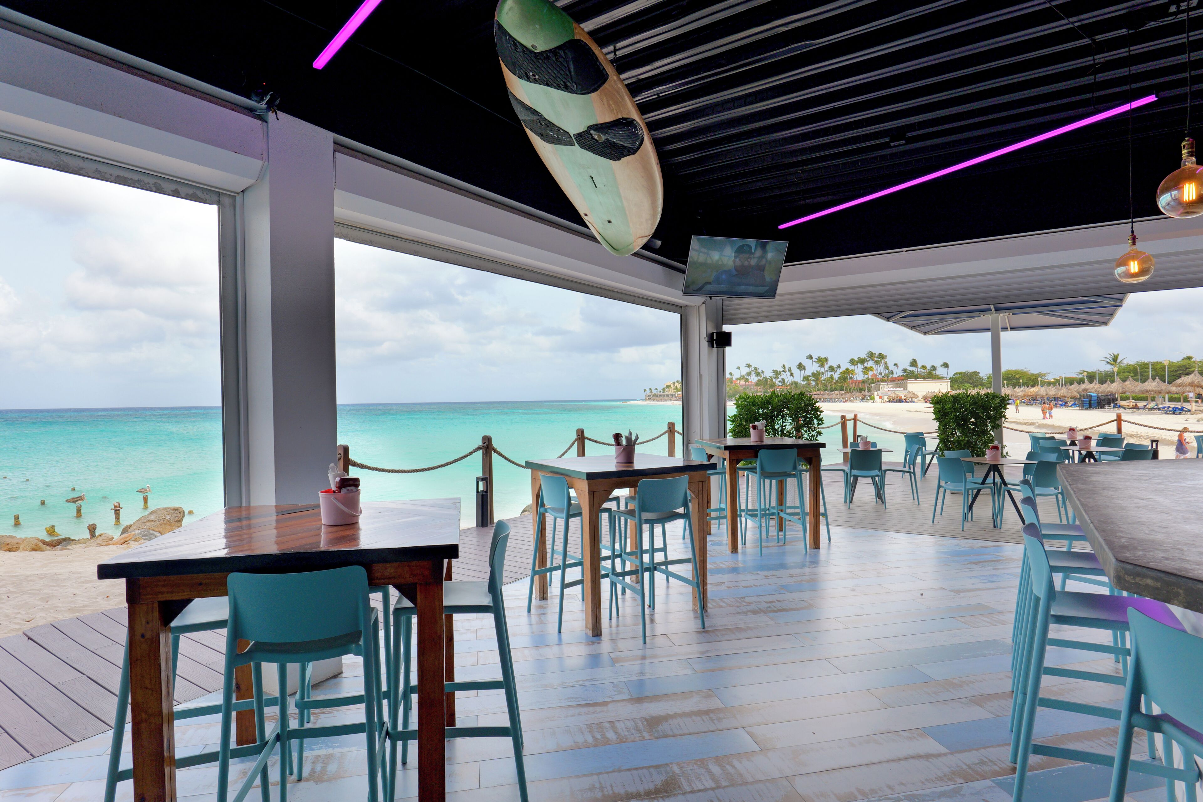 Un restaurante con vistas a la playa y al océano, con mesas y sillas de madera, una tabla de surf colgante y una pantalla de televisión.