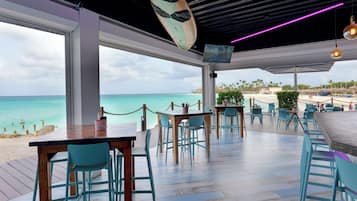 Un restaurante con vistas a la playa y al océano, con mesas y sillas de madera, una tabla de surf colgante y una pantalla de televisión.