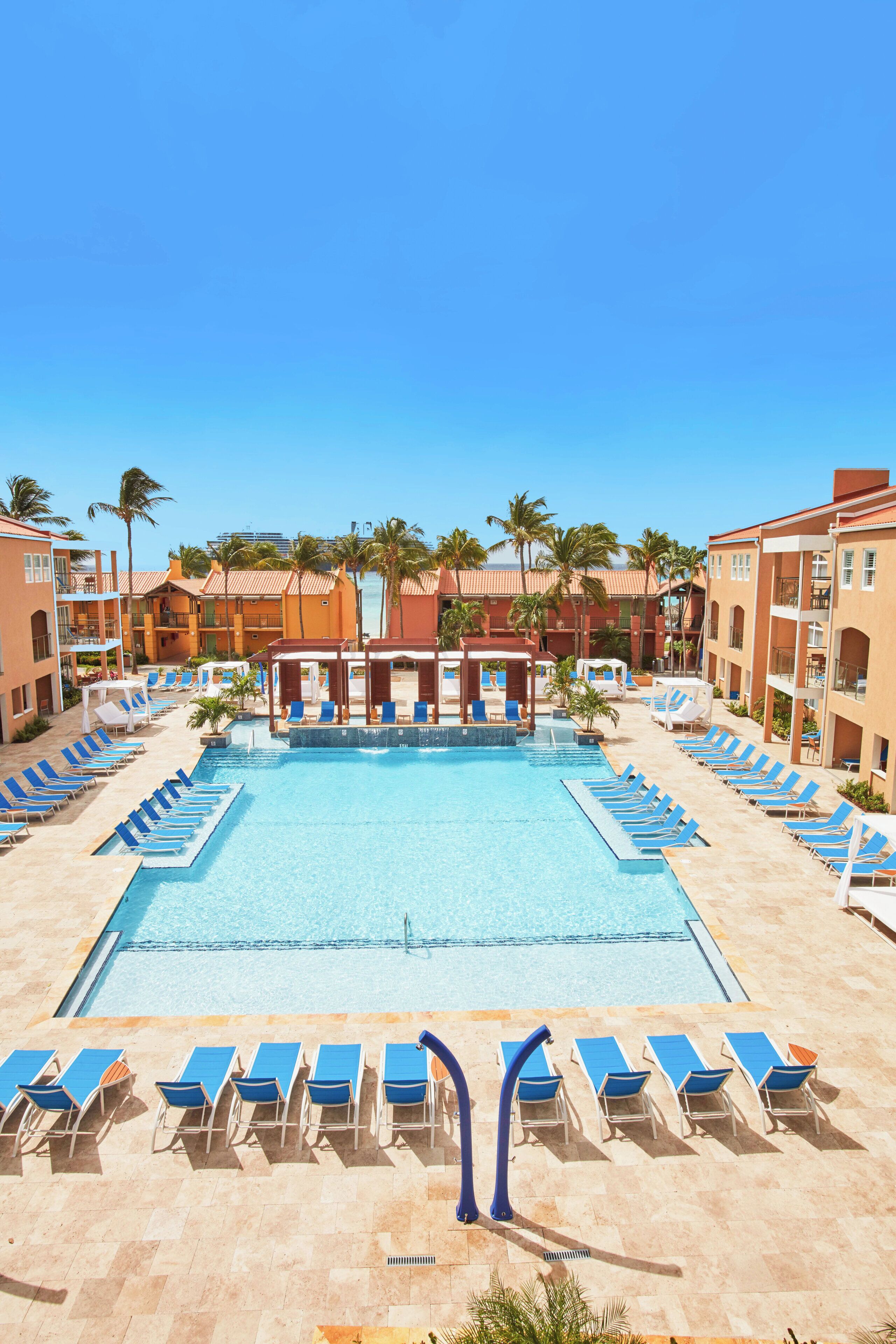Une piscine bordée de chaises longues bleues, de palmiers et d’un bâtiment doté de balcons.
