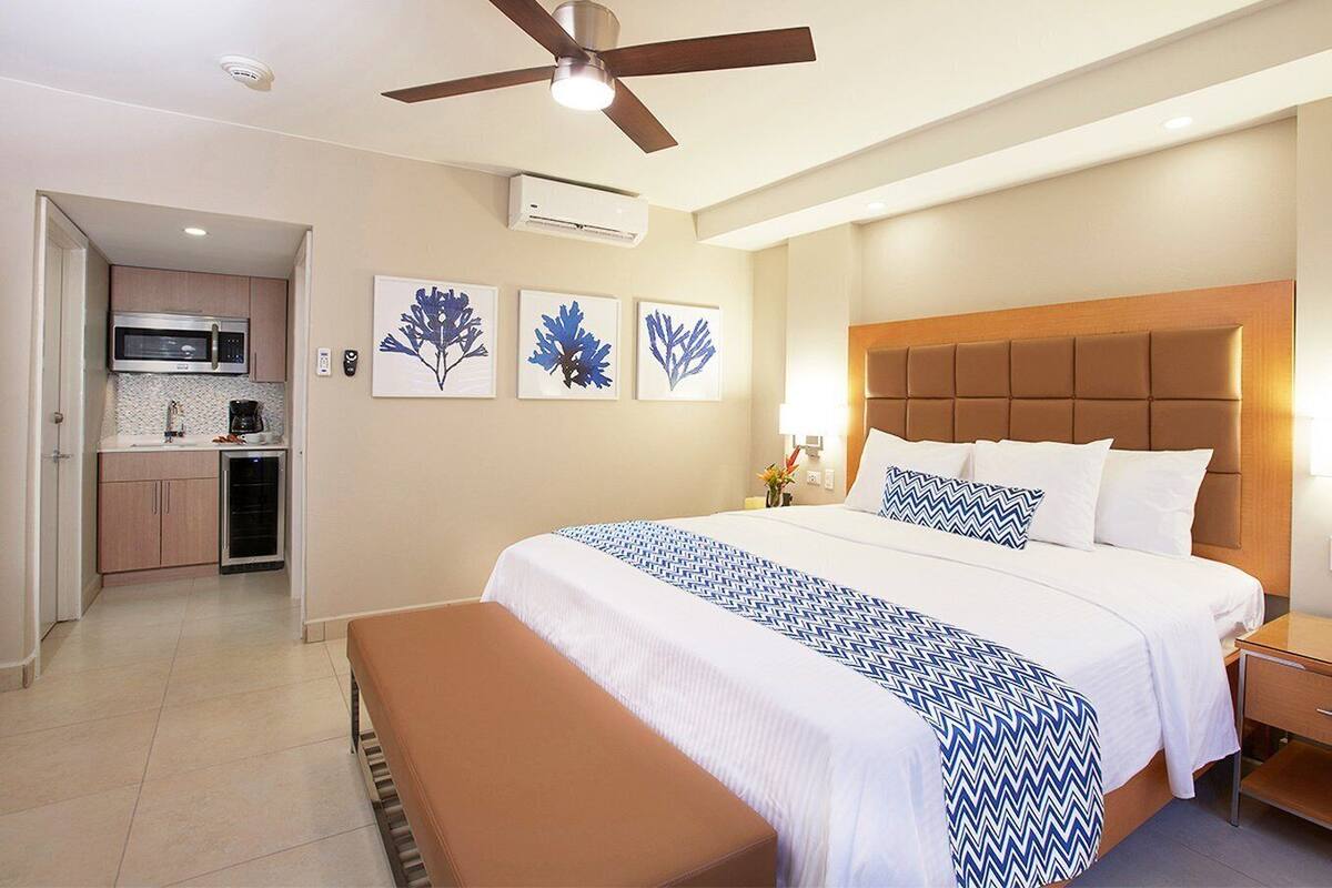 A hotel room with a bed, a bench, a ceiling fan, a kitchenette, and wall art.