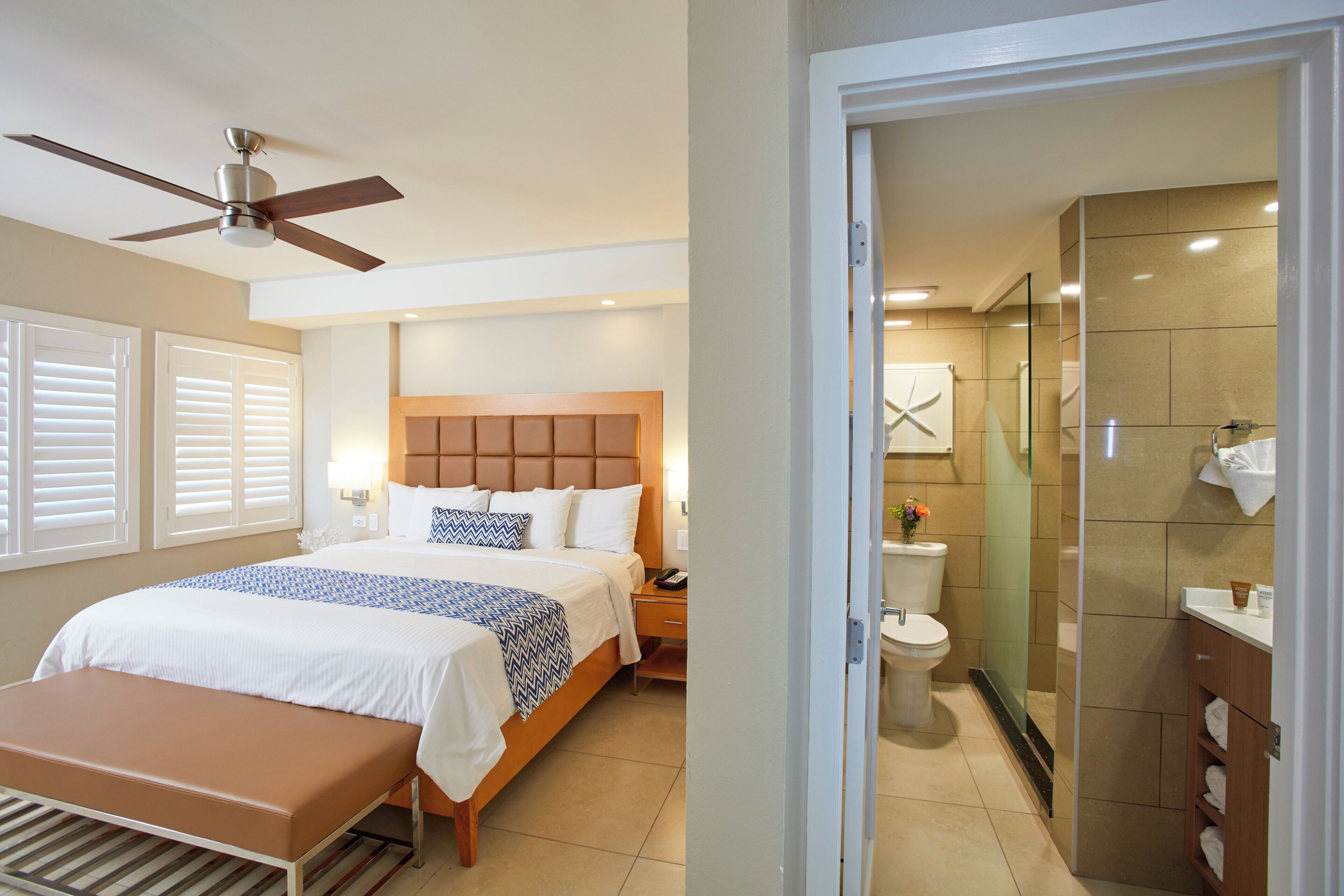 A hotel room with a bed, a bench, a ceiling fan, a nightstand, a bedside table, a bathroom with a toilet, a shower, and a glass door leading to the bathroom.