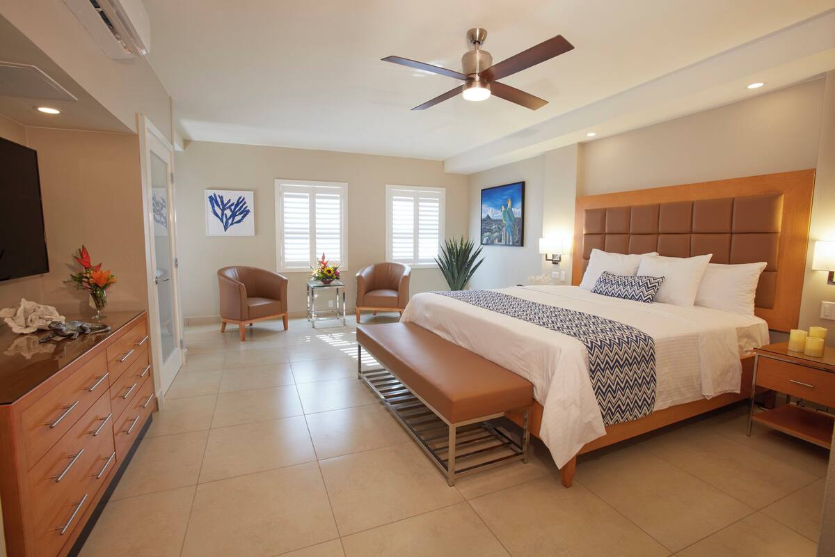 A bedroom with a bed, a bench, a desk, a chair, a TV, and a ceiling fan.