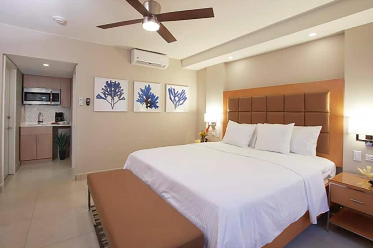 A hotel room with a bed, a nightstand, a bench, a ceiling fan, an air conditioning unit, and a kitchenette in the background.