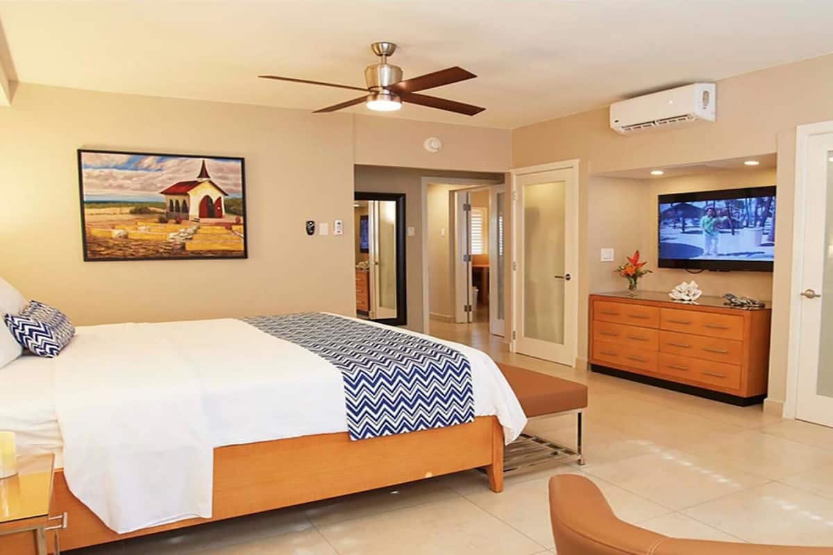 A hotel room with a bed, a ceiling fan, a TV, a dresser, and a painting on the wall.