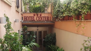 Entrada de un hotel con un letrero que dice "HOTEL CENTER1". Hay plantas en macetas y un pequeño jardín frente a la entrada.