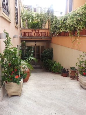 Entrada de un hotel con un letrero que dice "HOTEL CENTER1". Hay plantas en macetas y un pequeño jardín frente a la entrada.