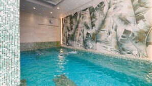 Una piscina interior con agua azul cristalina, rodeada por una pared de azulejos con un mural de hojas tropicales.
