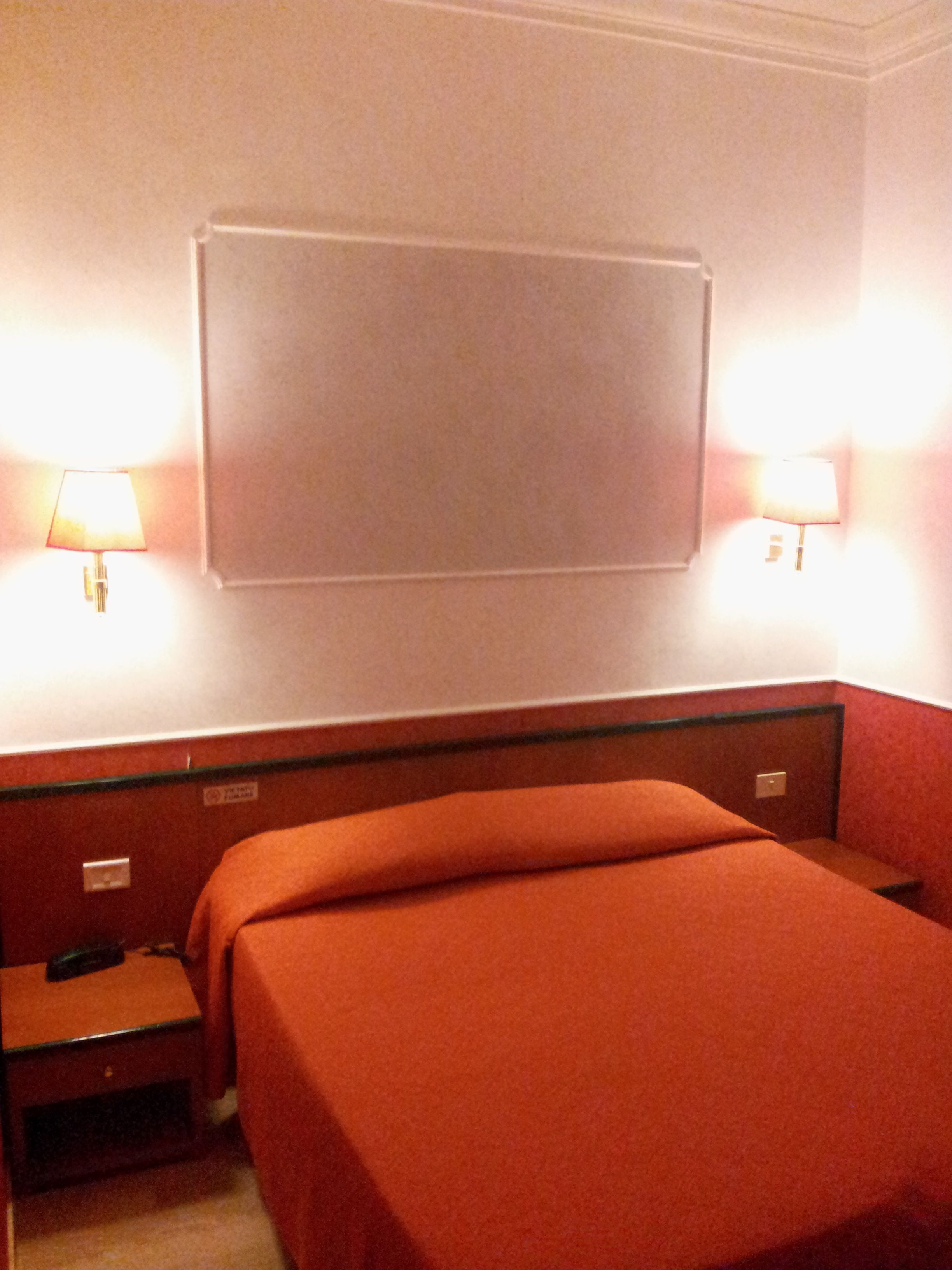 A hotel room with a bed, a nightstand, and two wall-mounted lamps.