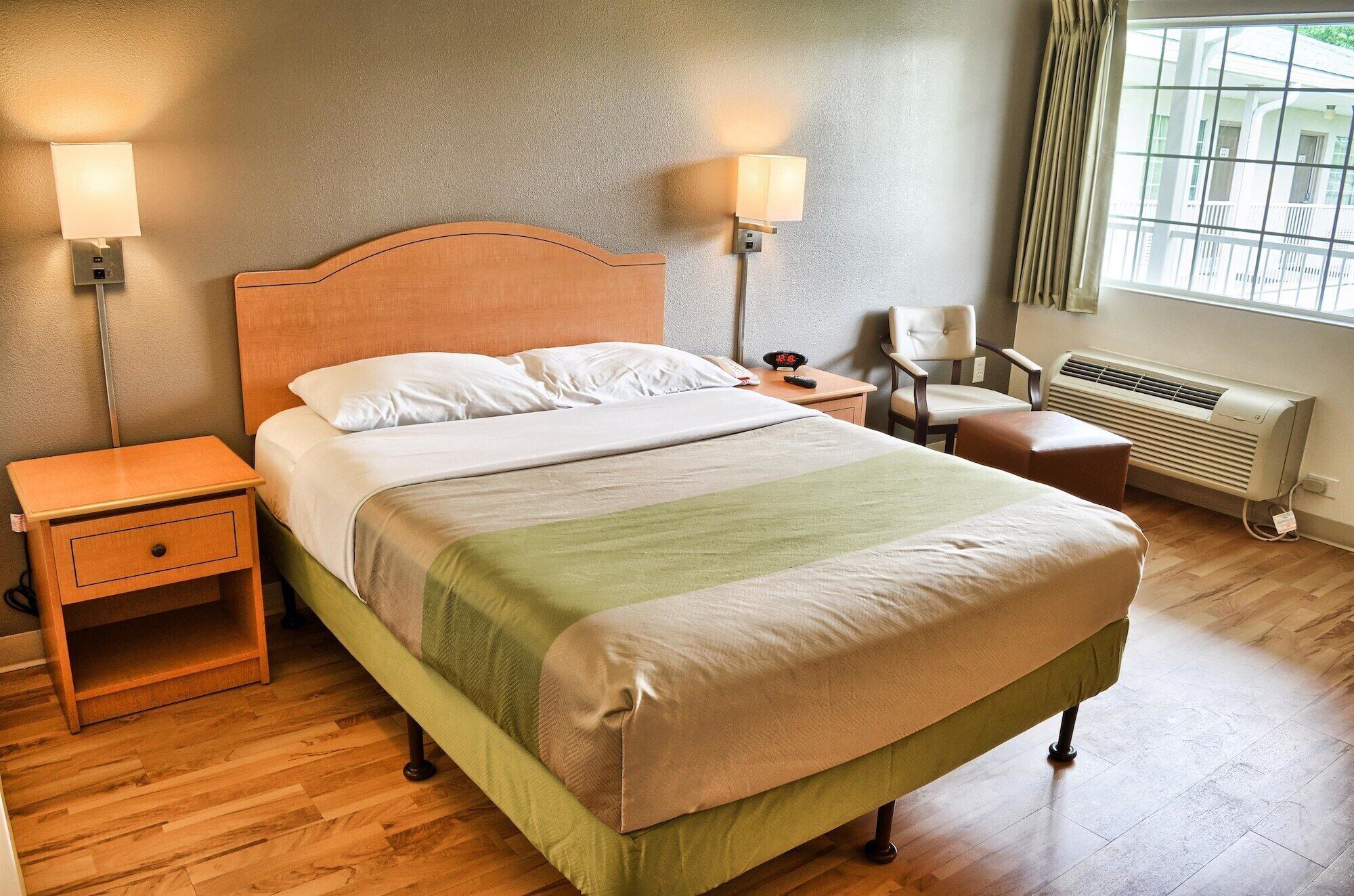 Una cama bien hecha con cabecera de madera, dos mesitas de noche, una silla, un calentador y una ventana con cortinas.