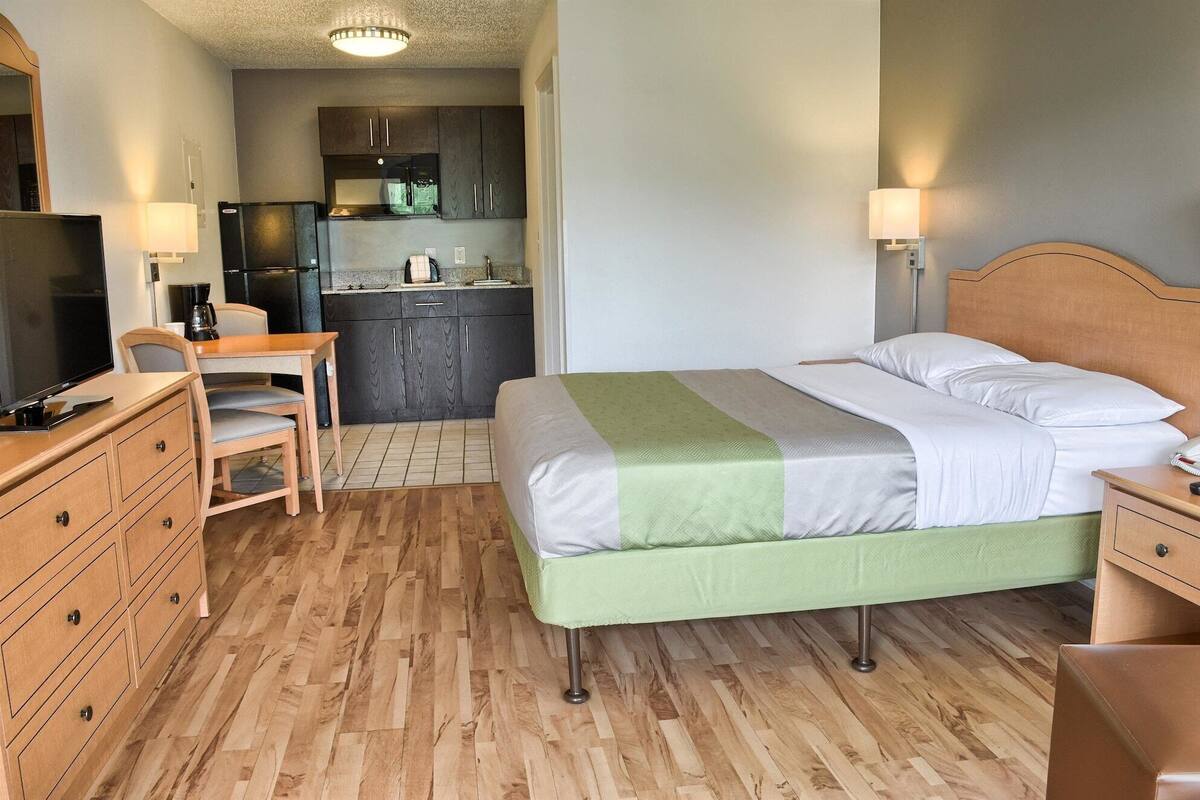 A compact hotel room with a bed, bedside table, TV, wooden dresser, kitchenette, and a small dining area.
