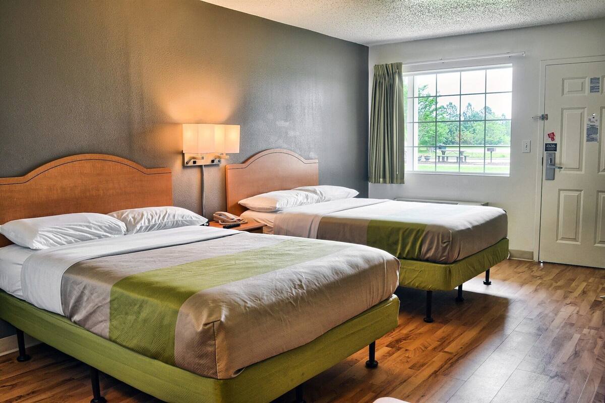 A hotel room with two beds, a nightstand with a lamp, a window with curtains, and a door with a lock.