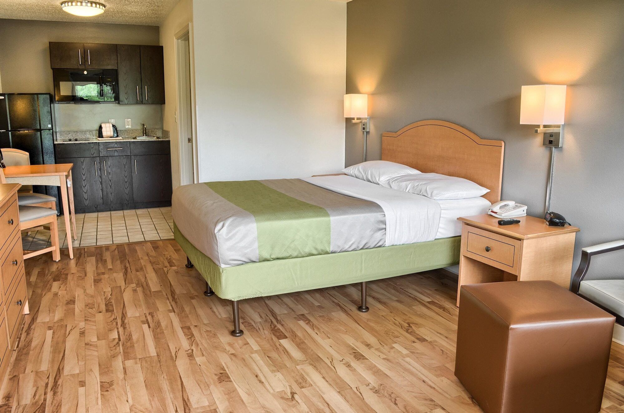 Quarto de hotel com cama, abajures, uma escrivaninha, uma cadeira e uma cozinha compacta ao fundo.