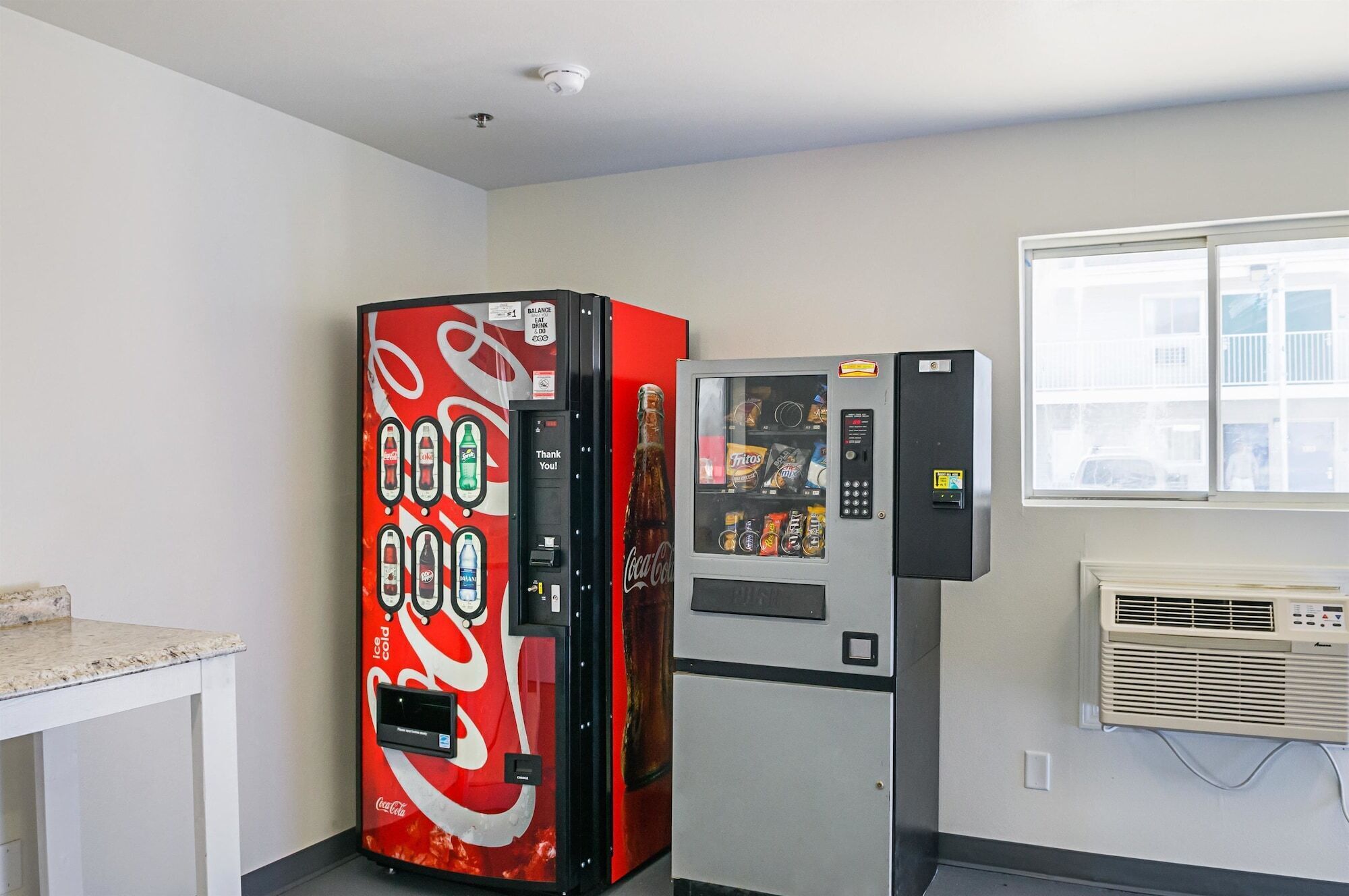Ein Zimmer mit einem Coca-Cola-Automaten, einer Minikühlschrank und einem Fenster mit Jalousien.