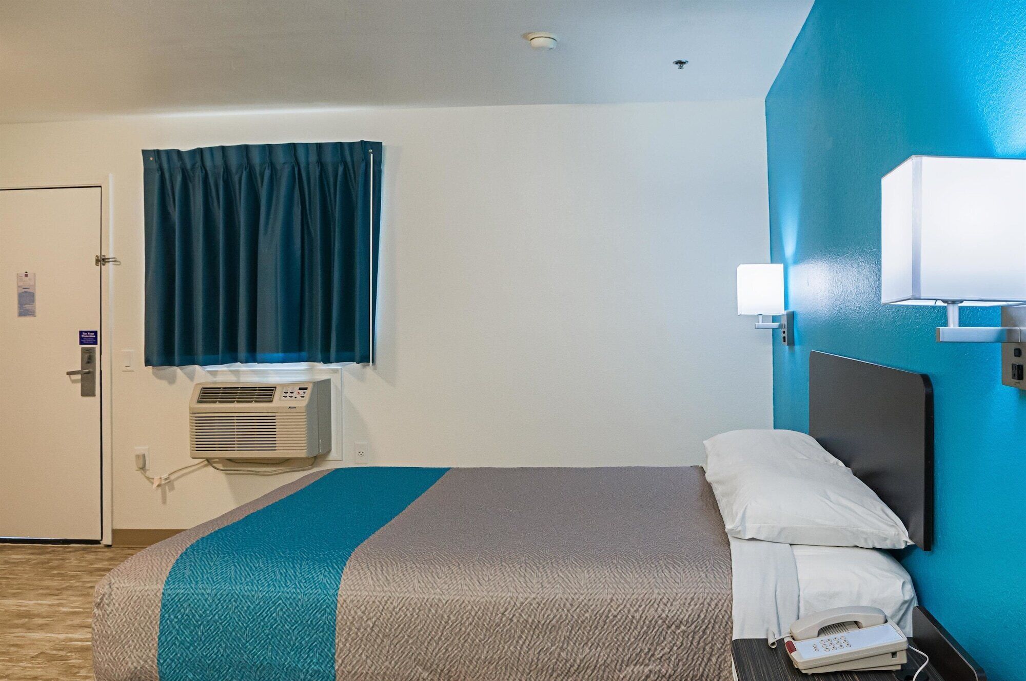Une chambre d’hôtel équipée d’un lit, d’un téléphone sur la table de chevet, d’une fenêtre avec des rideaux, d’un climatiseur et d’une lampe fixée au mur.