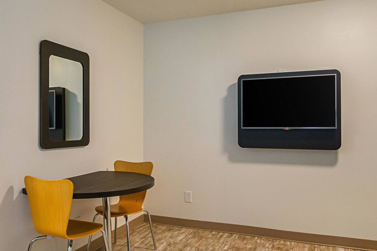 Ein Zimmer mit einem an der Wand befestigten Flachbildfernseher, einem kleinen runden Tisch mit zwei gelben Stühlen und einem Spiegel.