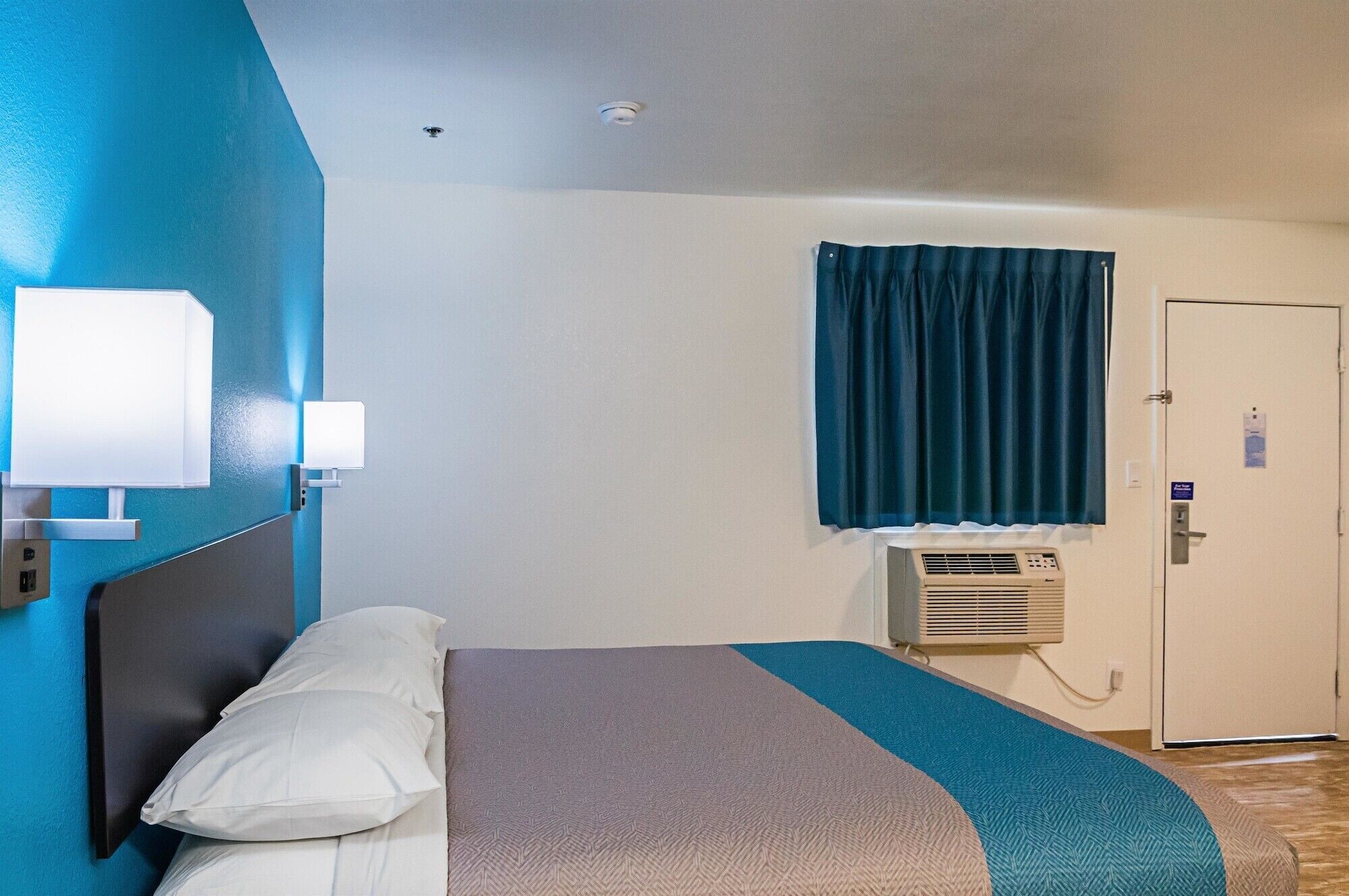 A hotel room with a bed, a wall-mounted air conditioner, a door with a sign, and a window with curtains.