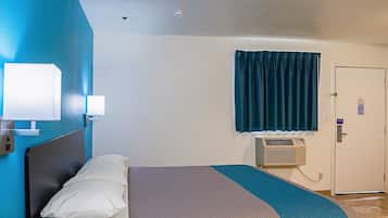 A hotel room with a bed, a wall-mounted air conditioner, a door with a sign, and a window with curtains.