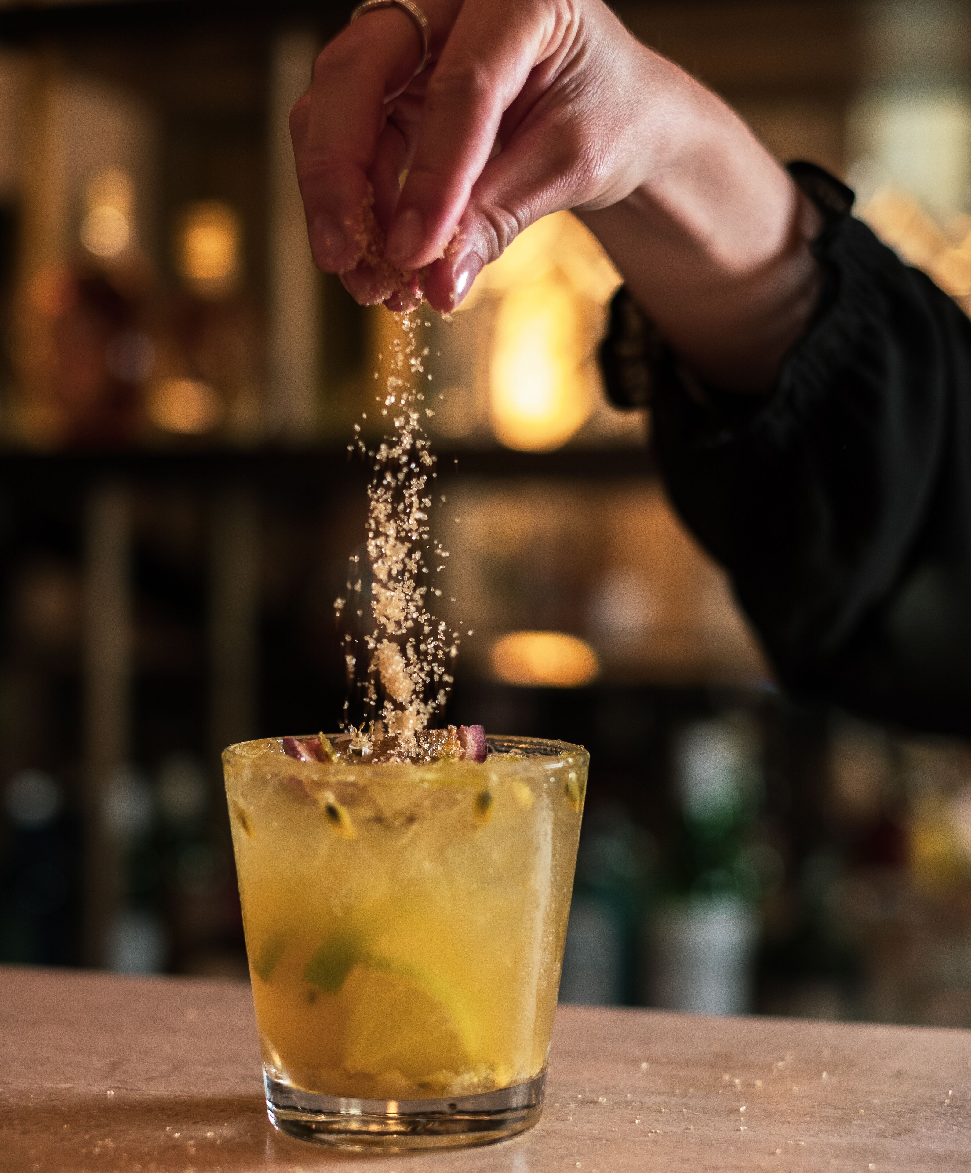 A hand sprinkling a granular substance into a glass containing a yellowish liquid with ice cubes.