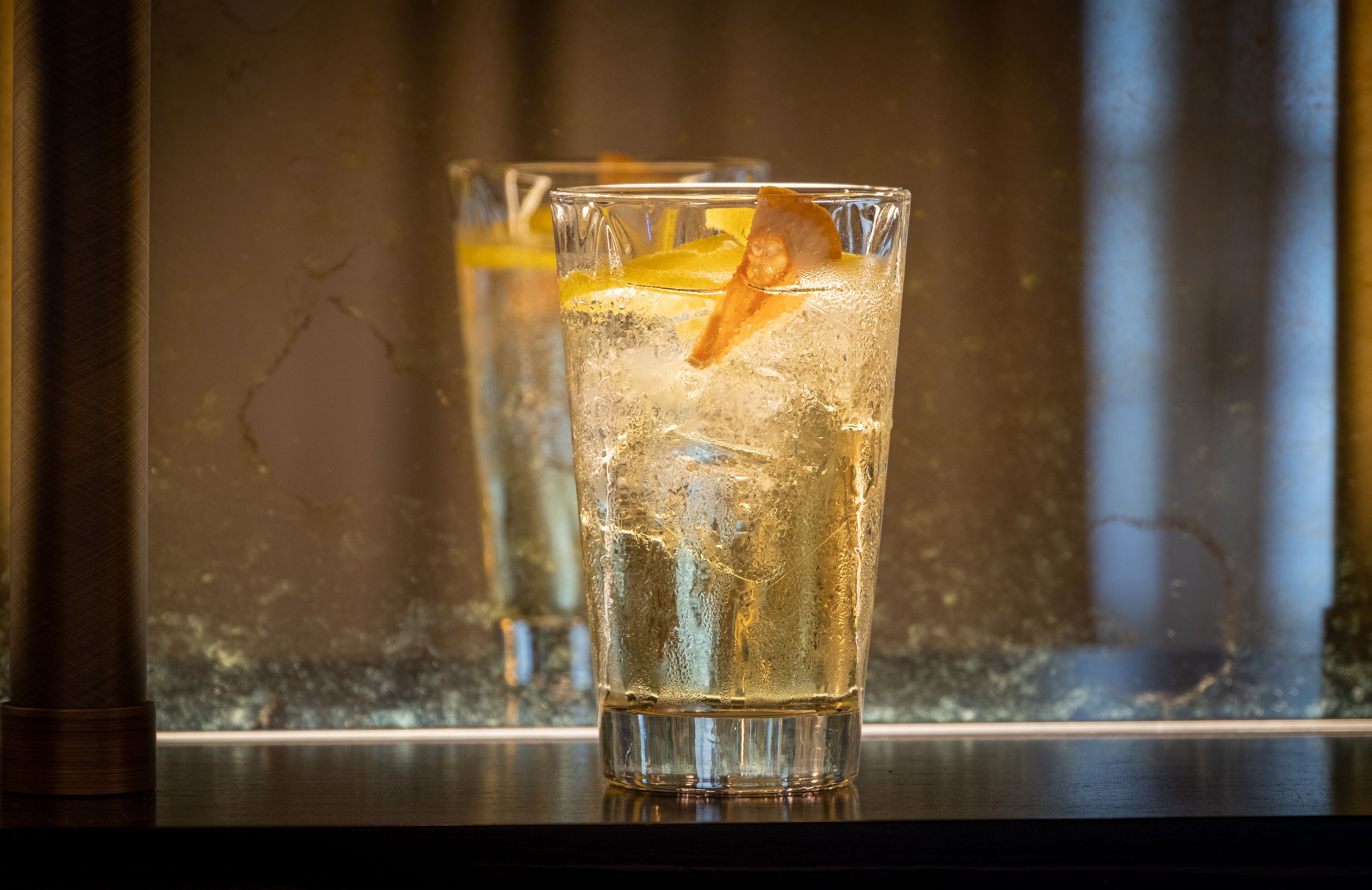 Deux verres contenant une boisson claire, avec des glaçons et une tranche d’orange en guise de décoration, posés sur une surface réfléchissante.