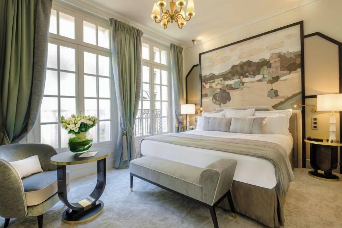 A hotel room with a large bed, a desk, a chair, a vase of flowers, and a painting on the wall.