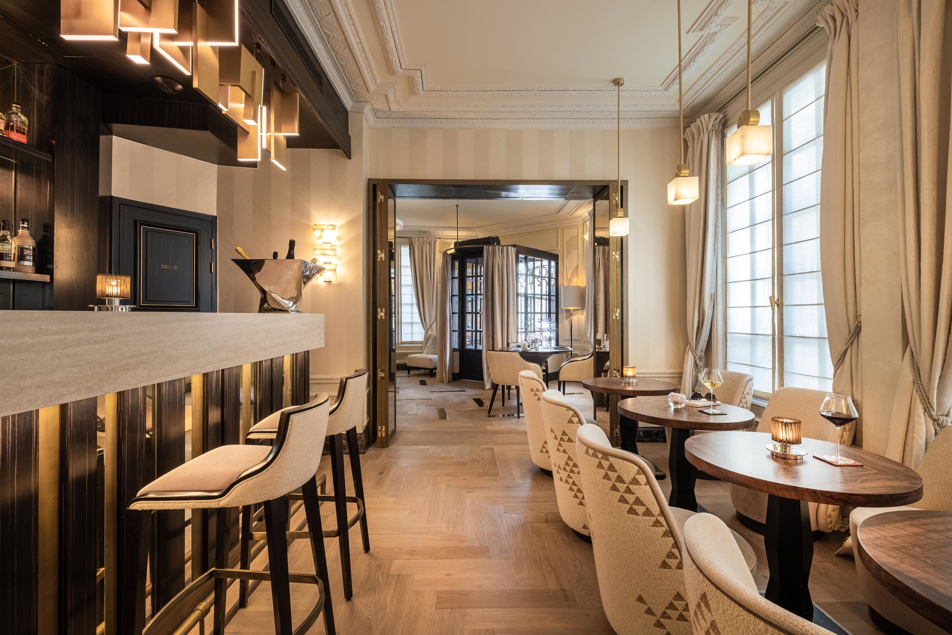 Un espace bar avec des tabourets, un comptoir et des bouteilles. On y trouve également des tables avec des chaises et, au fond, une salle à manger.