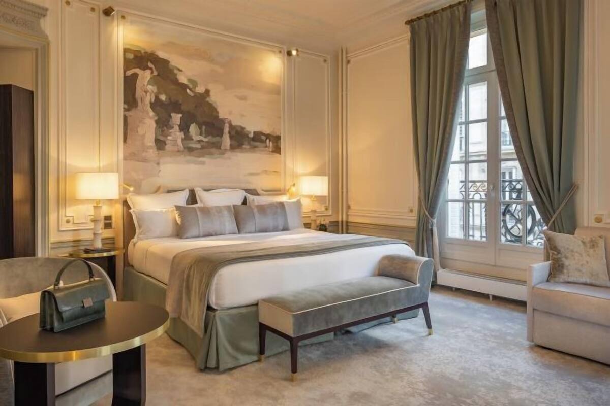 A hotel room with a large bed, two bedside tables with lamps, a bench, a sofa, and a large window with curtains.