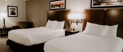 A hotel room with two beds, a chair, a nightstand with a lamp, and framed pictures on the wall.