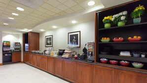 A hotel reception area with a long counter, various food and beverage stations, and a display of flowers and fruits.