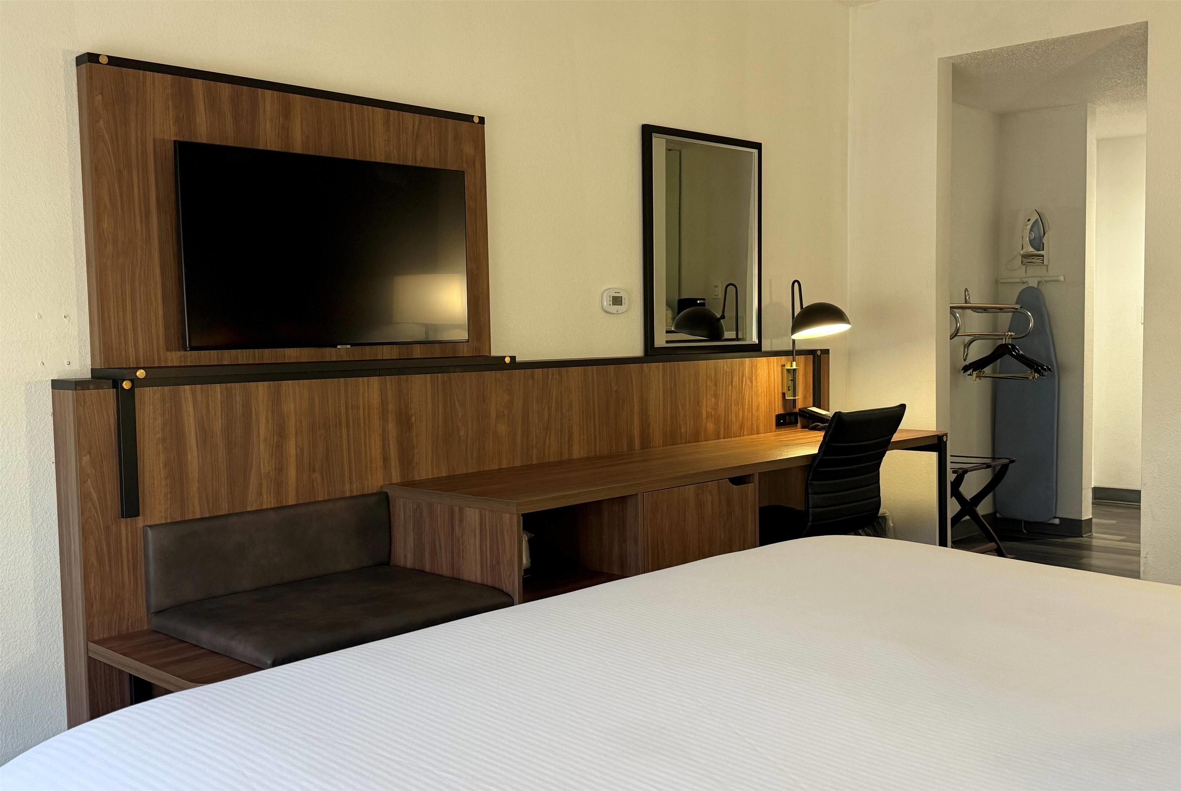 A hotel room with a bed, a wooden desk, a television, a mirror, and a lamp.