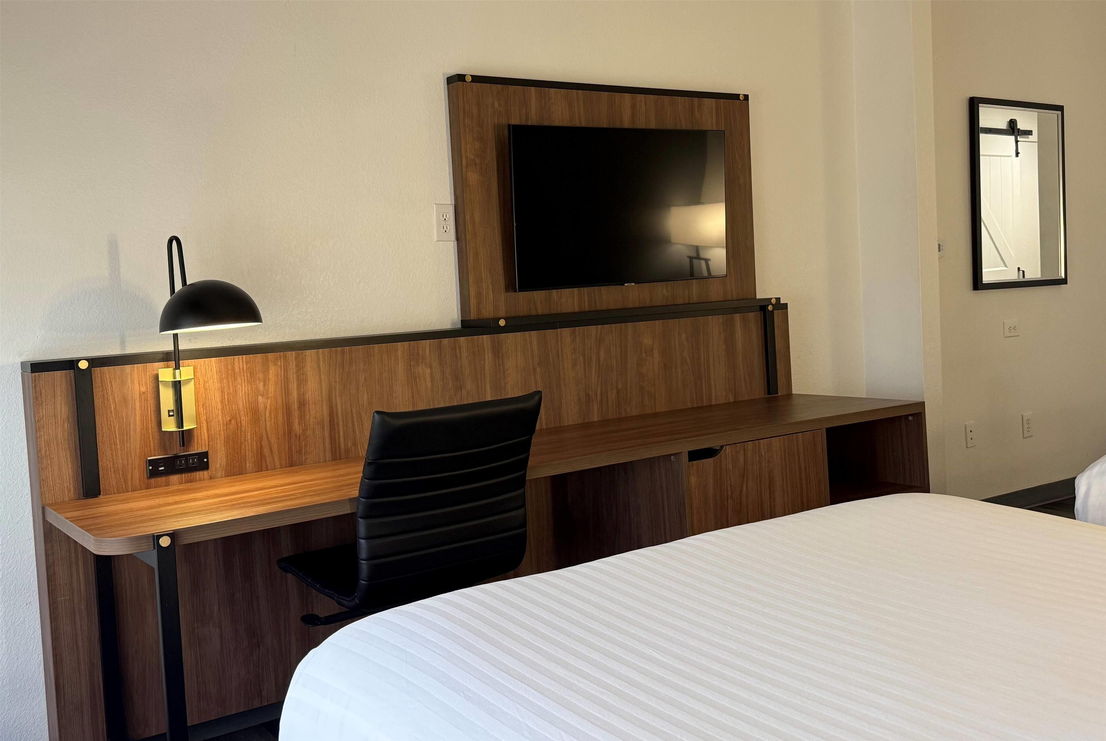 Een hotelkamer met een houten bureau, een zwarte stoel, een bed met wit beddegoed, een aan de muur bevestigde televisie en een spiegel.