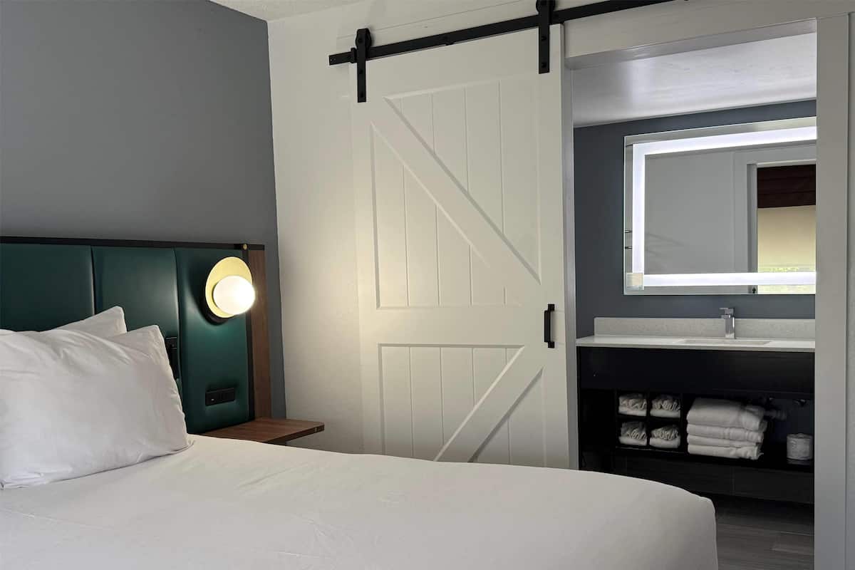 Una habitación de hotel con cama, cabecera con luz, puerta corredera estilo granero, baño con espejo y repisa con toallas.