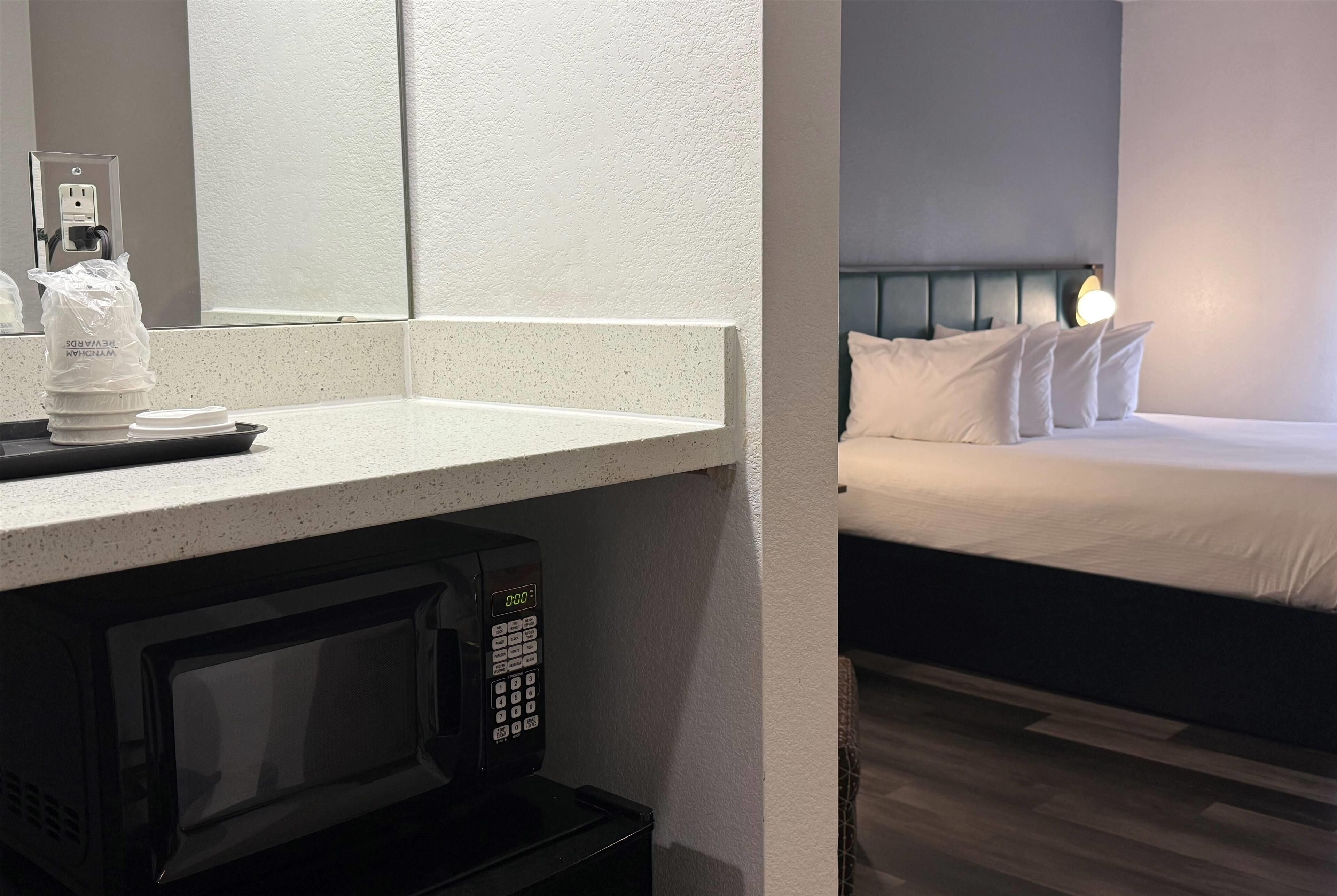 Quarto de hotel com micro-ondas na bancada, cama com roupa de cama branca e mesa de cabeceira com abajur.