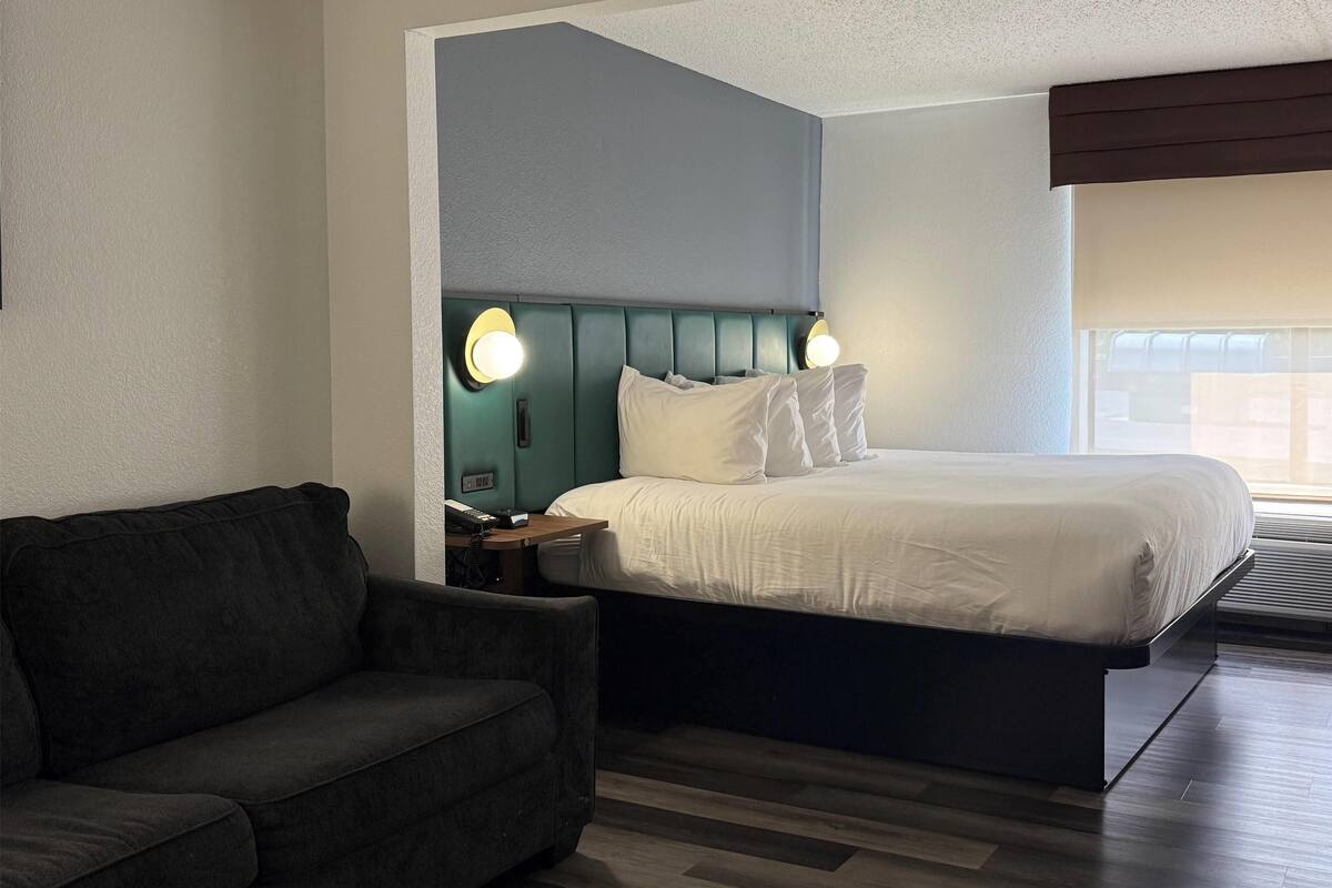 Una habitación de hotel moderna con una cama grande, un sofá de color oscuro y una mesita de noche con una lámpara.