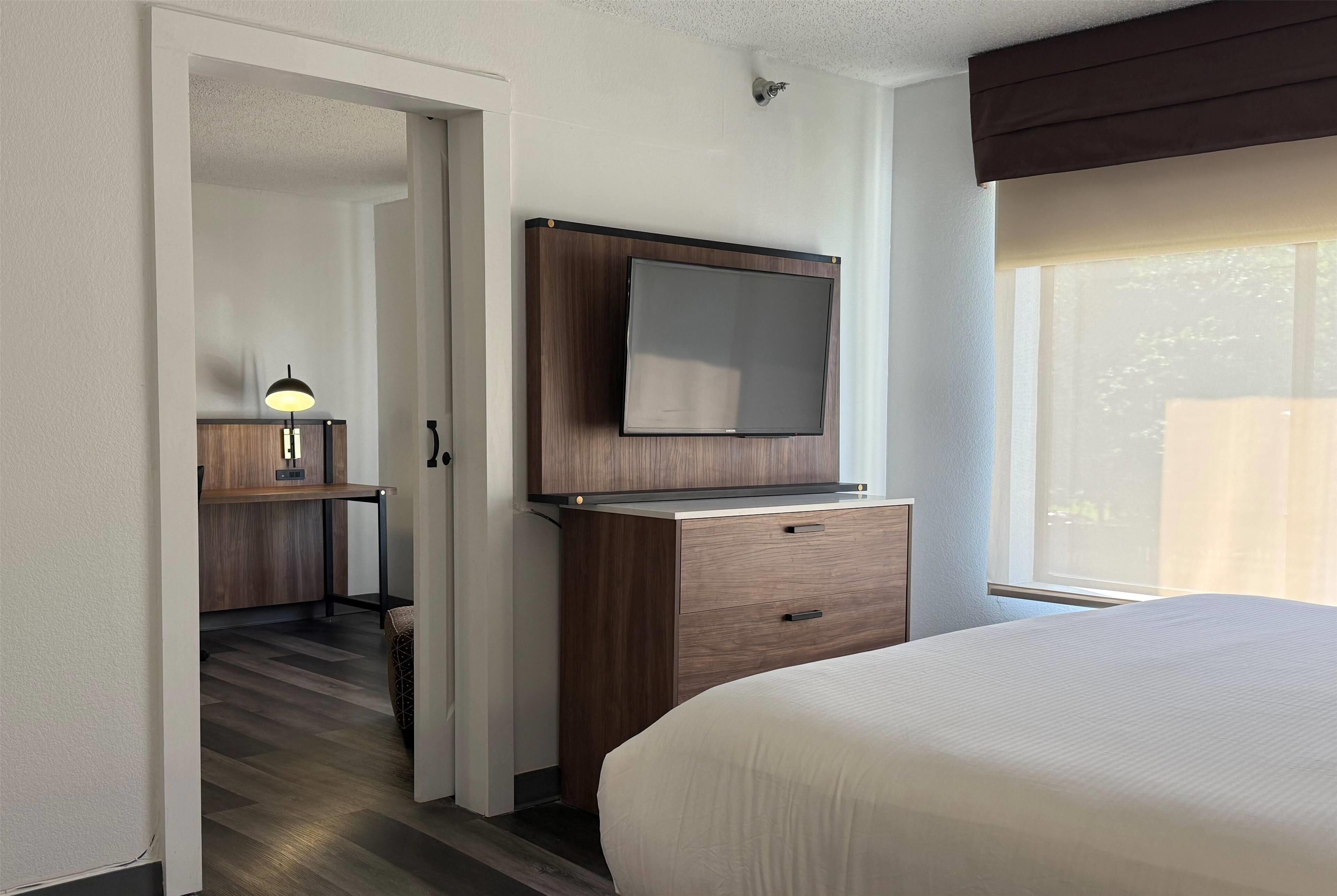A hotel room with a bed, a desk with a lamp, a TV mounted on a wooden cabinet, and a window with natural light.