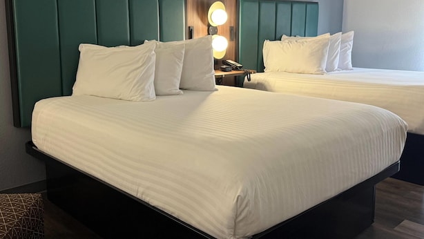 Quarto de hotel com duas camas, cada uma com roupa de cama branca e cabeceira verde-água. Há uma mesinha de cabeceira com luminária e telefone.