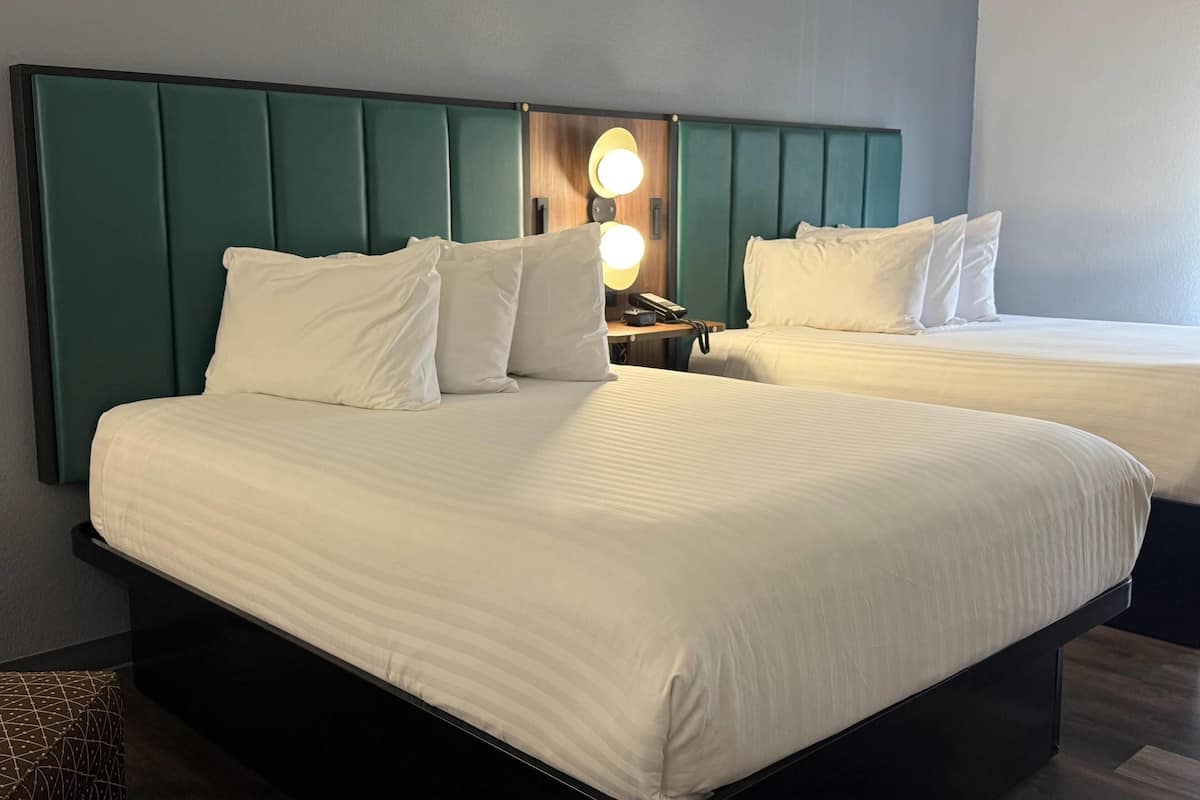 Habitación de hotel con dos camas, cada una con ropa blanca y cabeceros color turquesa. Hay una mesita de noche con una lámpara y un teléfono.