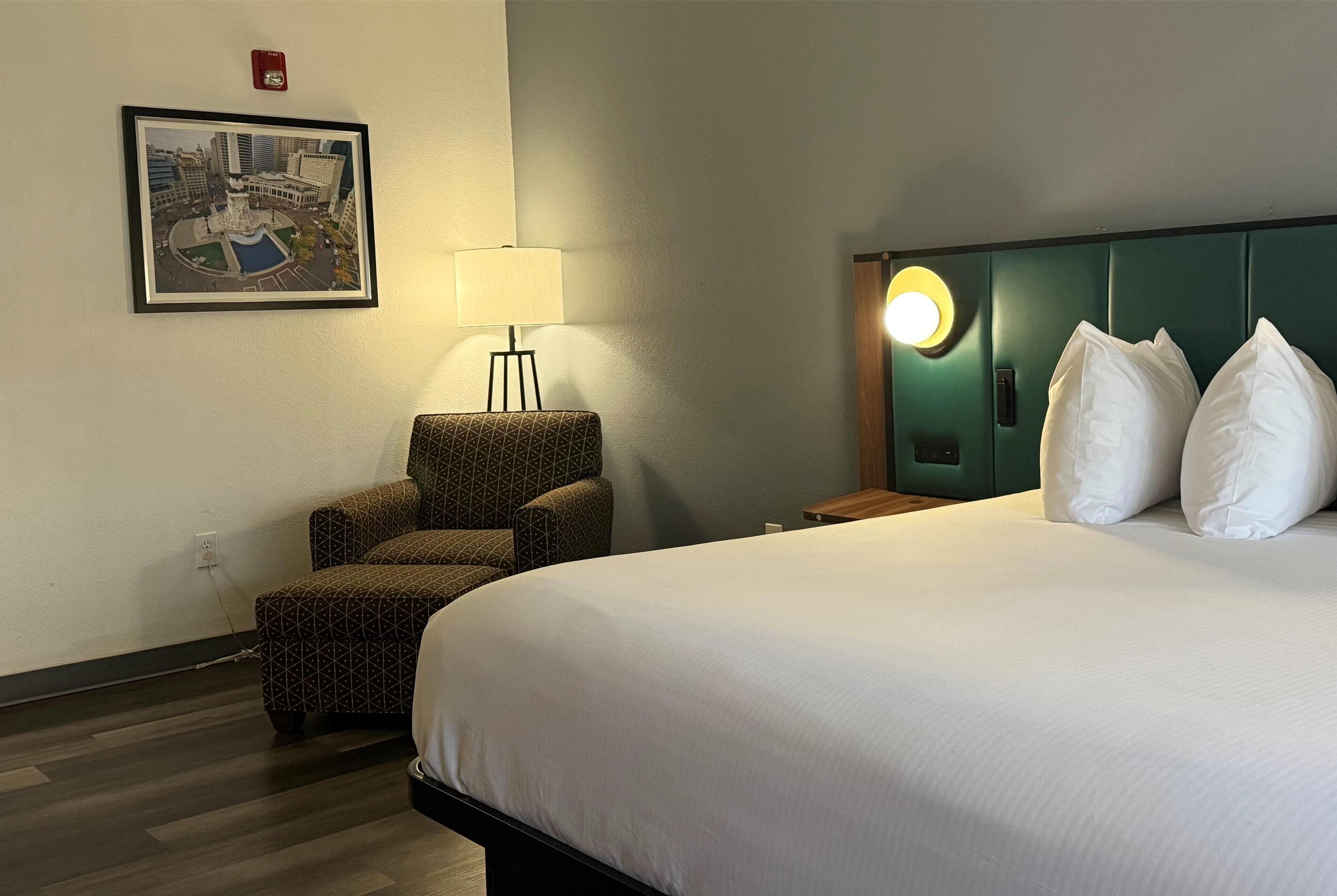 Quarto de hotel com uma cama grande, uma poltrona, um abajur, um quadro na parede e uma mesa de cabeceira com abajur.