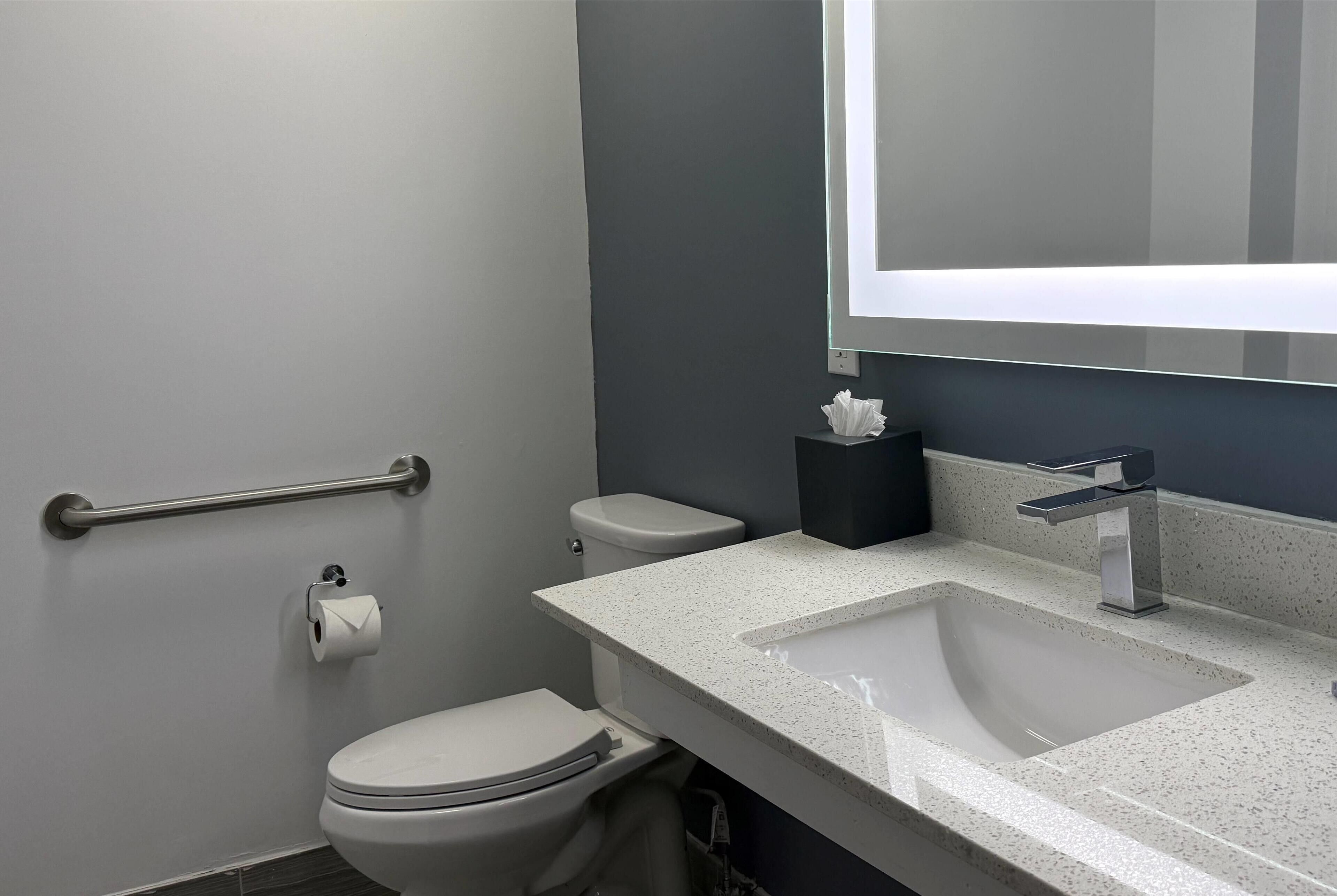 A modern bathroom with a white toilet, a sink with a mirror above it, and a handrail on the wall.