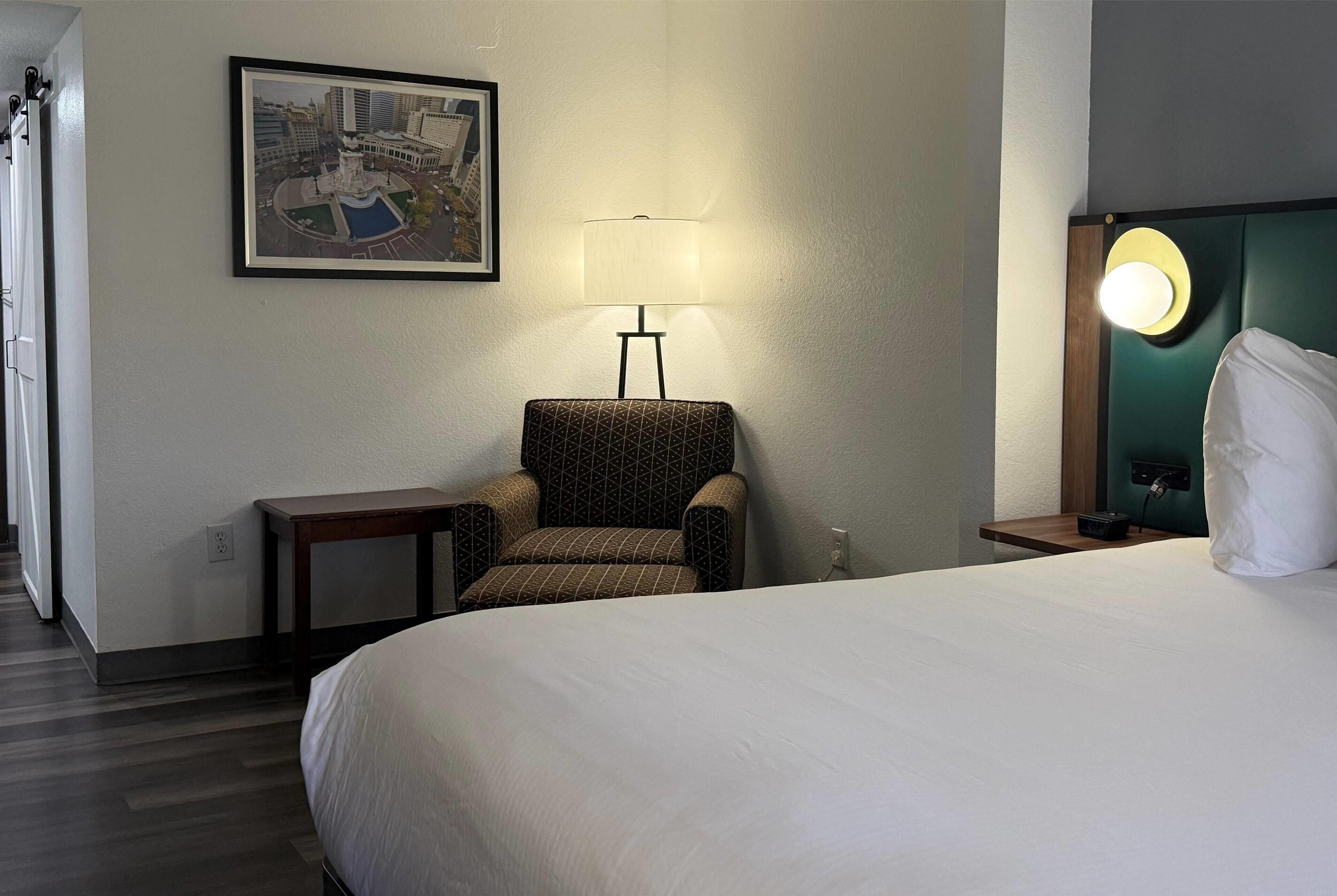 A hotel room with a large bed, a chair, a table, a lamp, and a framed picture on the wall.