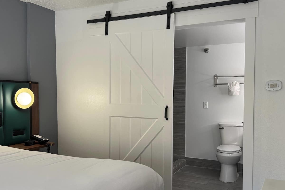 Una habitación de hotel con una cama, una mesita de noche con una lámpara, una puerta corrediza al baño y un termostato en la pared.