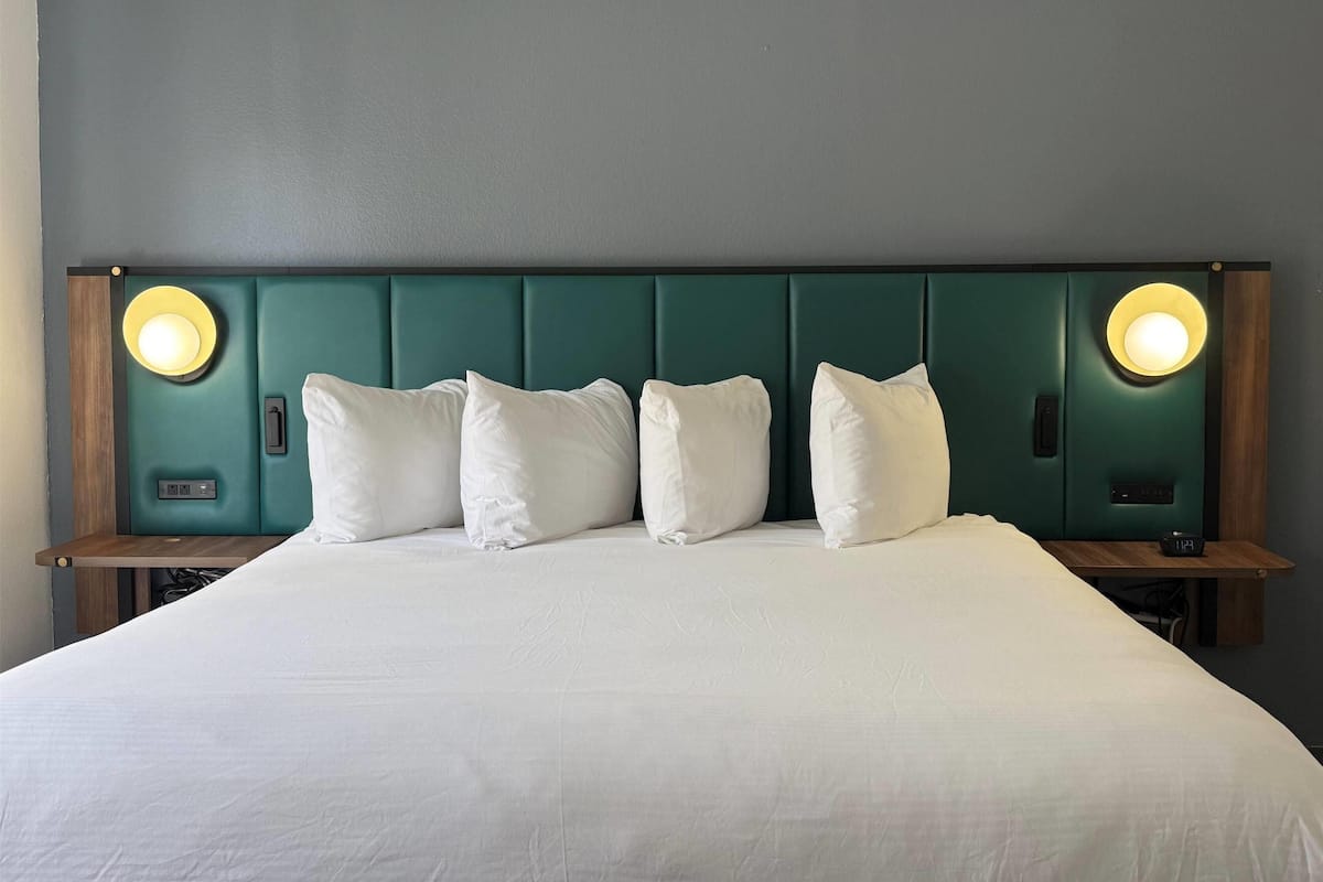 Una cama bien hecha con sábanas y almohadas blancas, un cabecero tapizado en color aguamarina con iluminación integrada y una mesita de noche con un reloj.