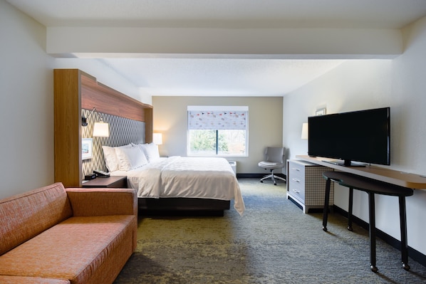 Ein Hotelzimmer mit Bett, Schreibtisch mit Fernseher, Sessel, Sofa und Fenster mit Jalousien.
