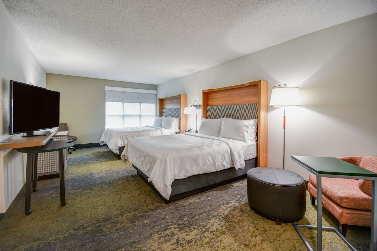 A hotel room with two beds, a desk with a flat-screen TV, a chair, a lamp, and a view of the outdoors.